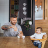 tattooed dad sitting at dining table pouring expressed breast milk into baby bottle from the Pumpd soft silicone breast pump while baby boy sits next to him in high chair smiling